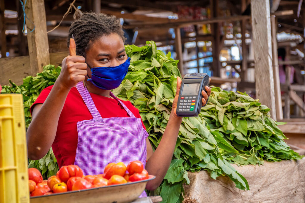 A female market vendor in Nigeria smiles while holding a POS machine, ready to accept a cashless payment for her fresh produce.